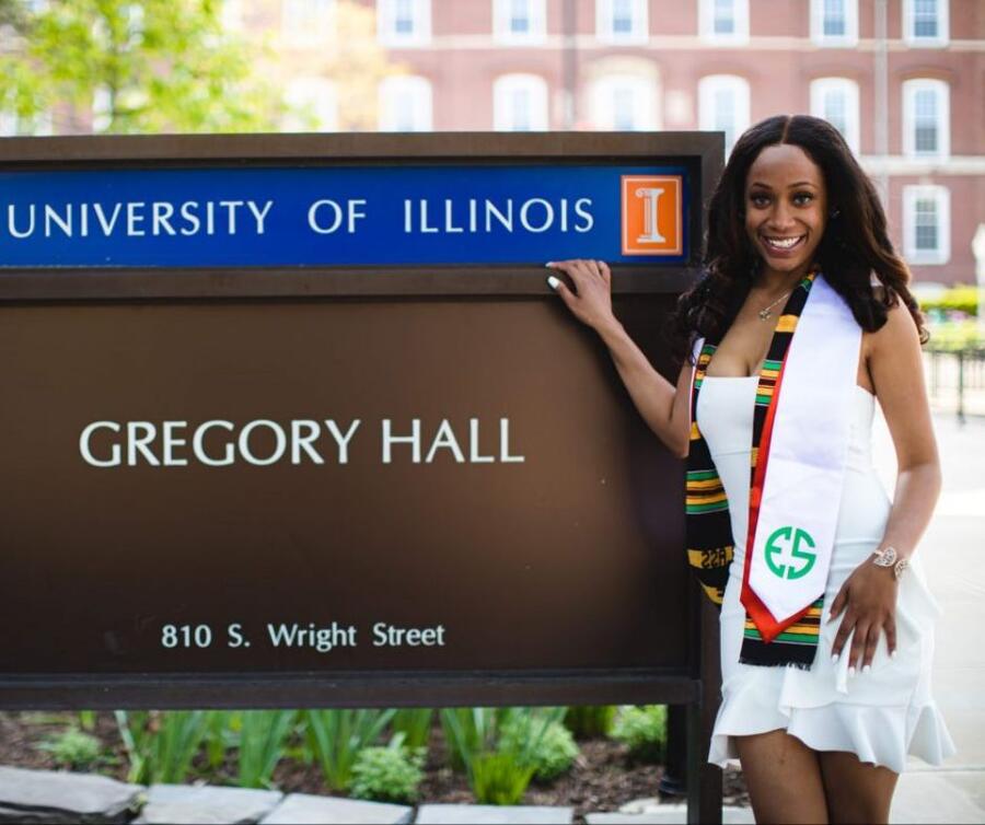 A student wearing a graduation sash stands before the Gregory Hall signboard, sporting a smile and posing with one hand placed on the sign.