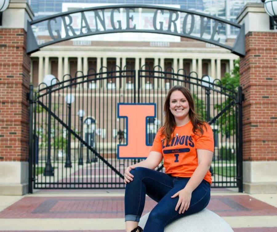 A student sits in front of the Grange Grove gate, wearing a smile and placing one hand on top of their knee.