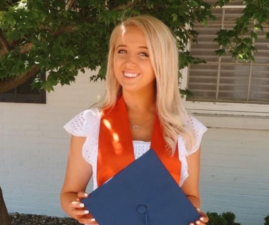 A student with a cheerful expression, wearing a graduation sash and holding a graduation cap in her hand.