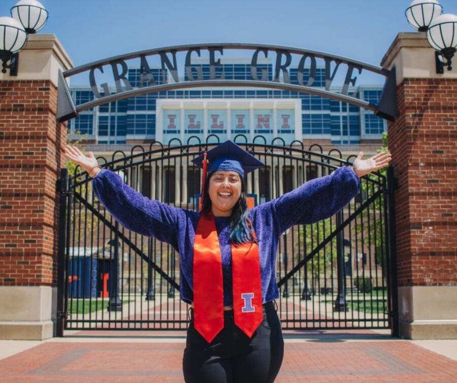 A jubilant student, dressed in a graduation sash and cap, stands with arms outstretched in a triumphant pose in front of the Grange Grove gate.