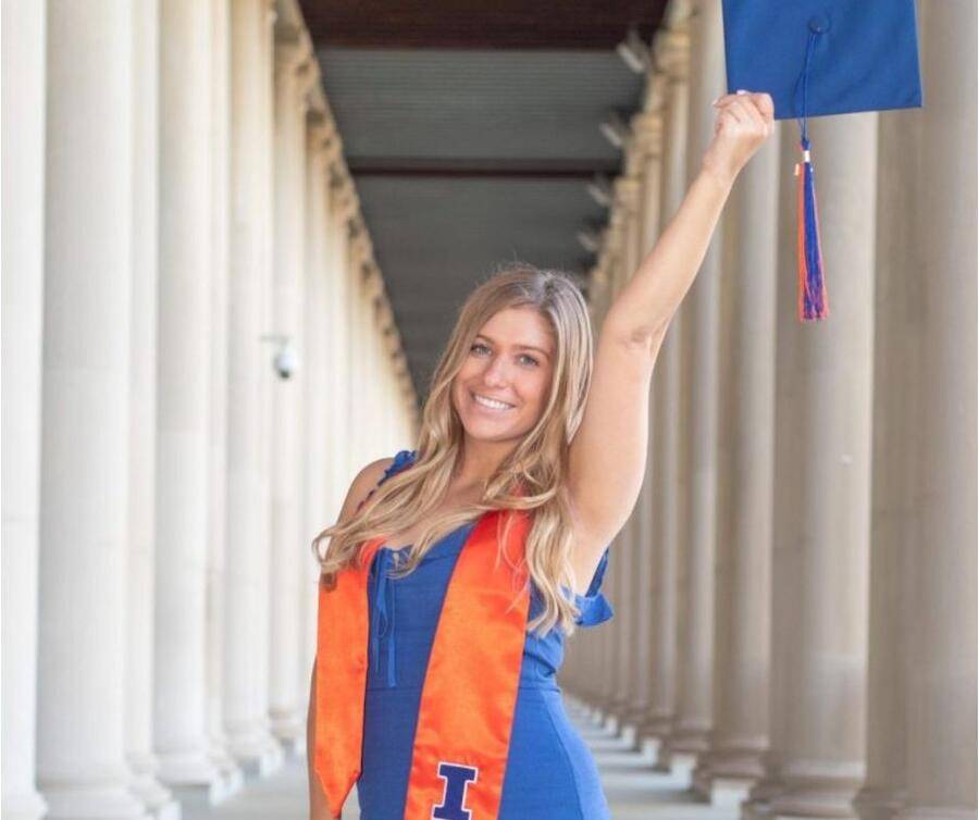 A cheerful student dressed in a blue gown and adorned with a graduation sash, while holding their cap aloft in one hand.