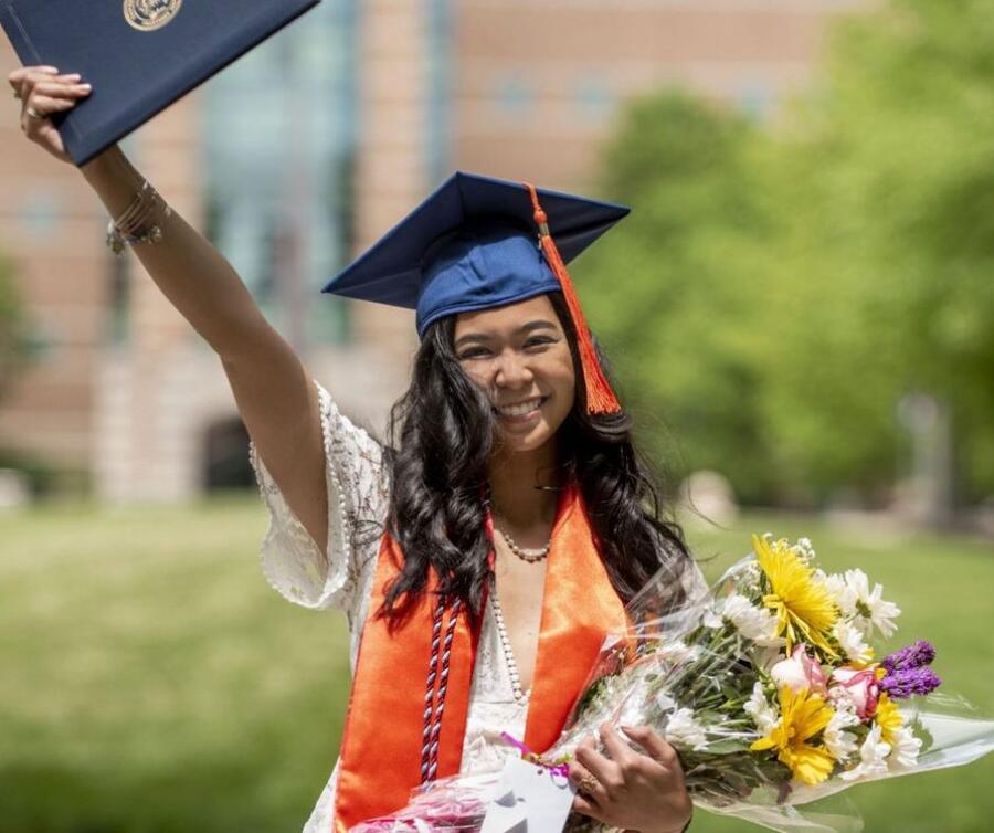 A jubilant student, adorned in a graduation sash and cap, lifts one hand high, clutching a degree, while holding a bouquet of flowers in the other, all accompanied by a warm smile.