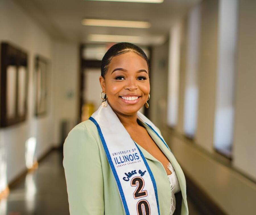 In a building's hallway, a student adorned with a graduation sash stands, radiating a joyful smile.