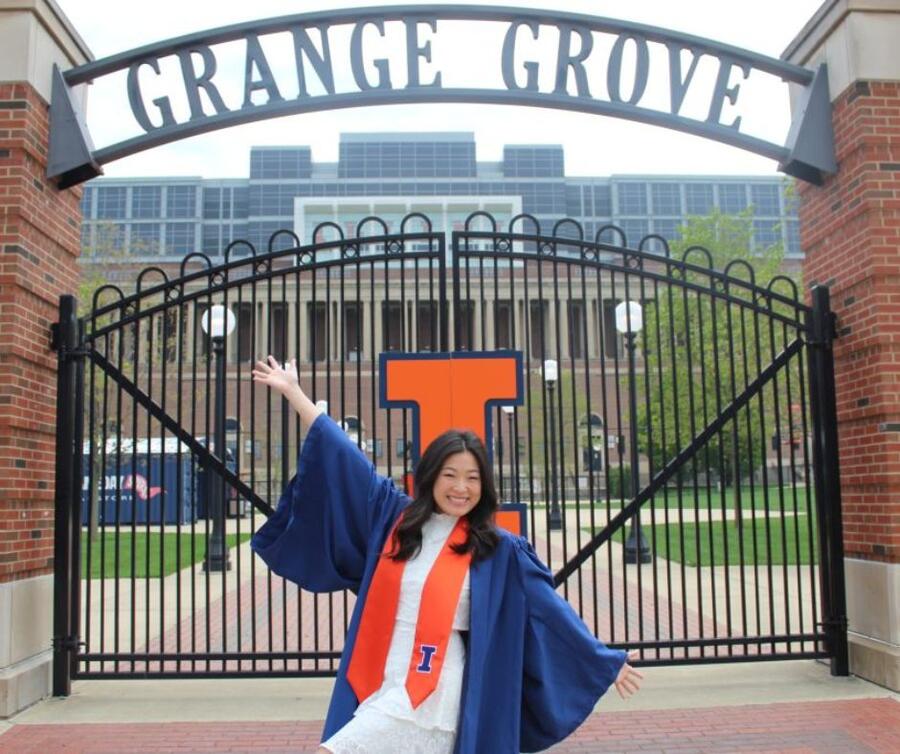 A student wearing a graduation gown and sash, with a big smile on their face, stands in front of the gate to the Grange Grove building, spreading their arms in a striking pose.