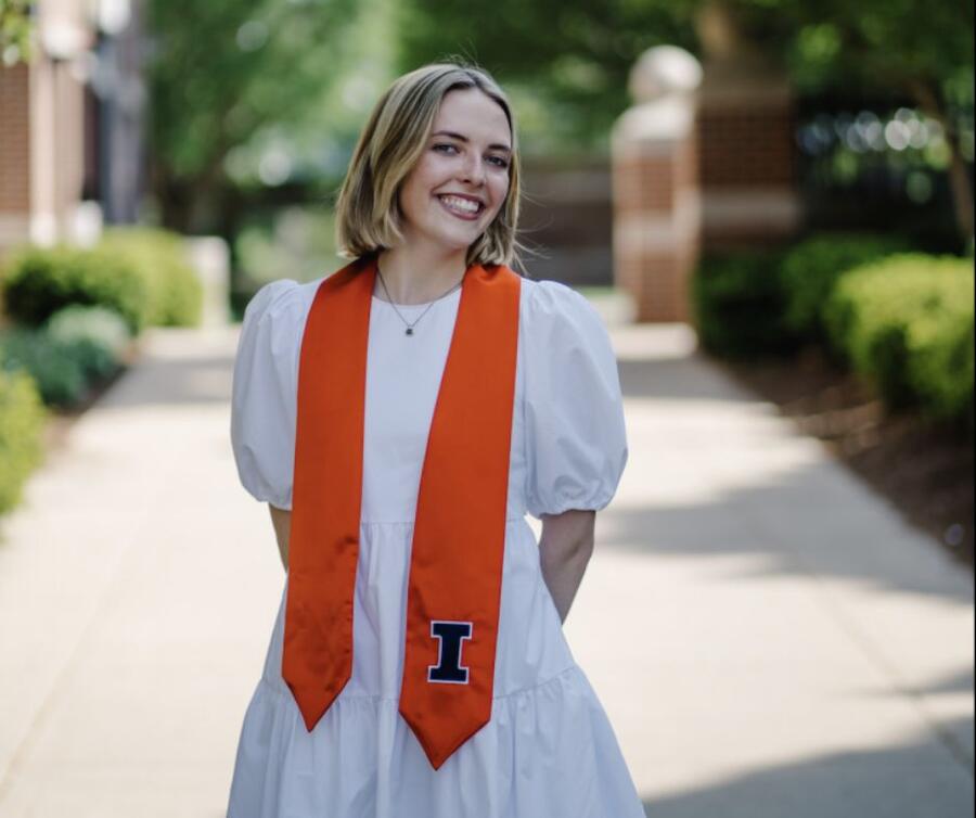 A student wearing a graduation sash stands joyfully in the middle of a pathway.